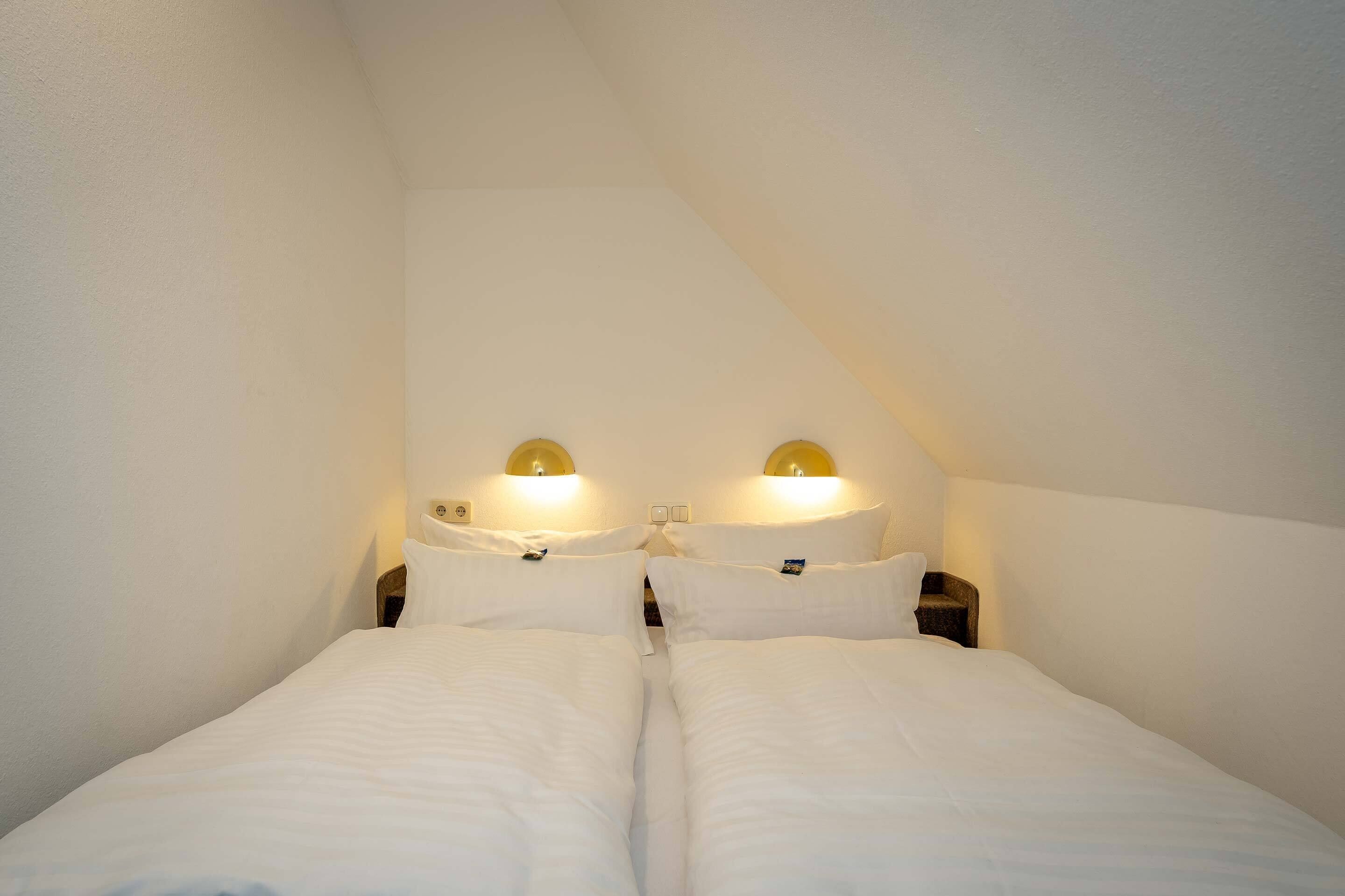 Une chambre double avec deux lits, chacun doté d’une tête de lit et de tables de chevet. Des lampes murales sont fixées de chaque côté des lits.