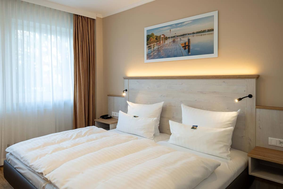 A hotel room with a large bed, a nightstand, a painting of a lakeside scene, and sheer curtains.