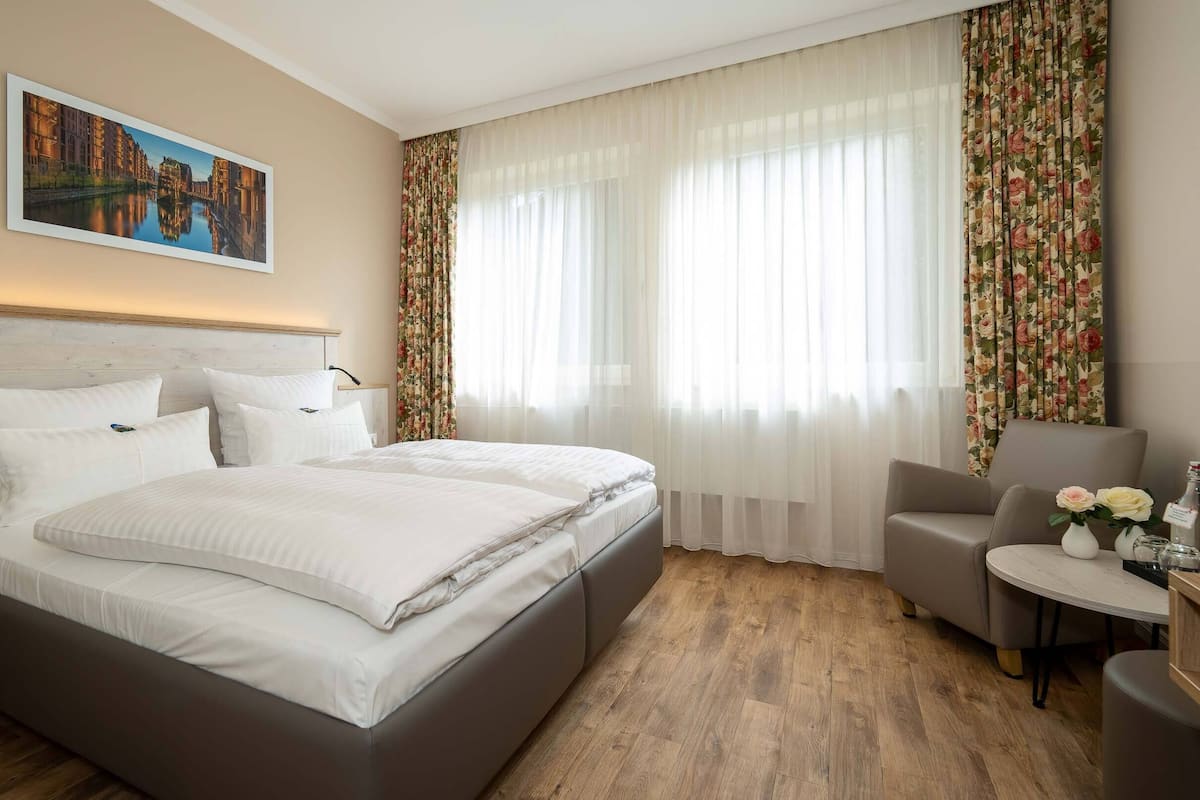 A hotel room with a large bed, a chair, a small table, and a painting on the wall.