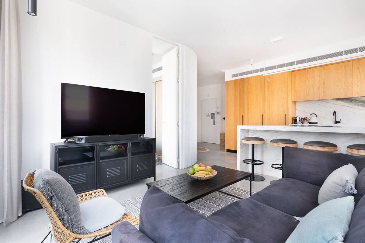 Ein modernes Wohnzimmer mit Fernseher, einer Couch, einem Couchtisch mit Früchten und einem Korbstuhl.