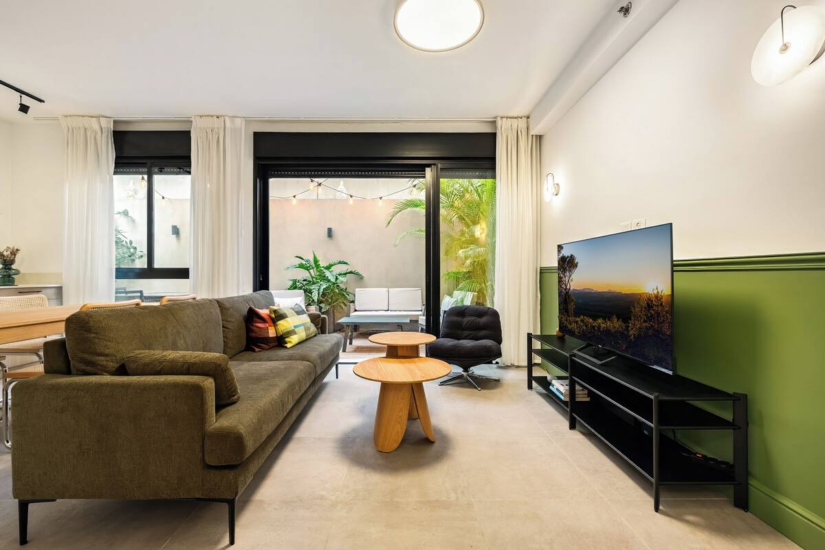 Ein modernes Wohnzimmer mit einer Couch, zwei runden Holztischen, einem schwarzen Sessel, einem Fernseher und einem Balkon mit Pflanzen.