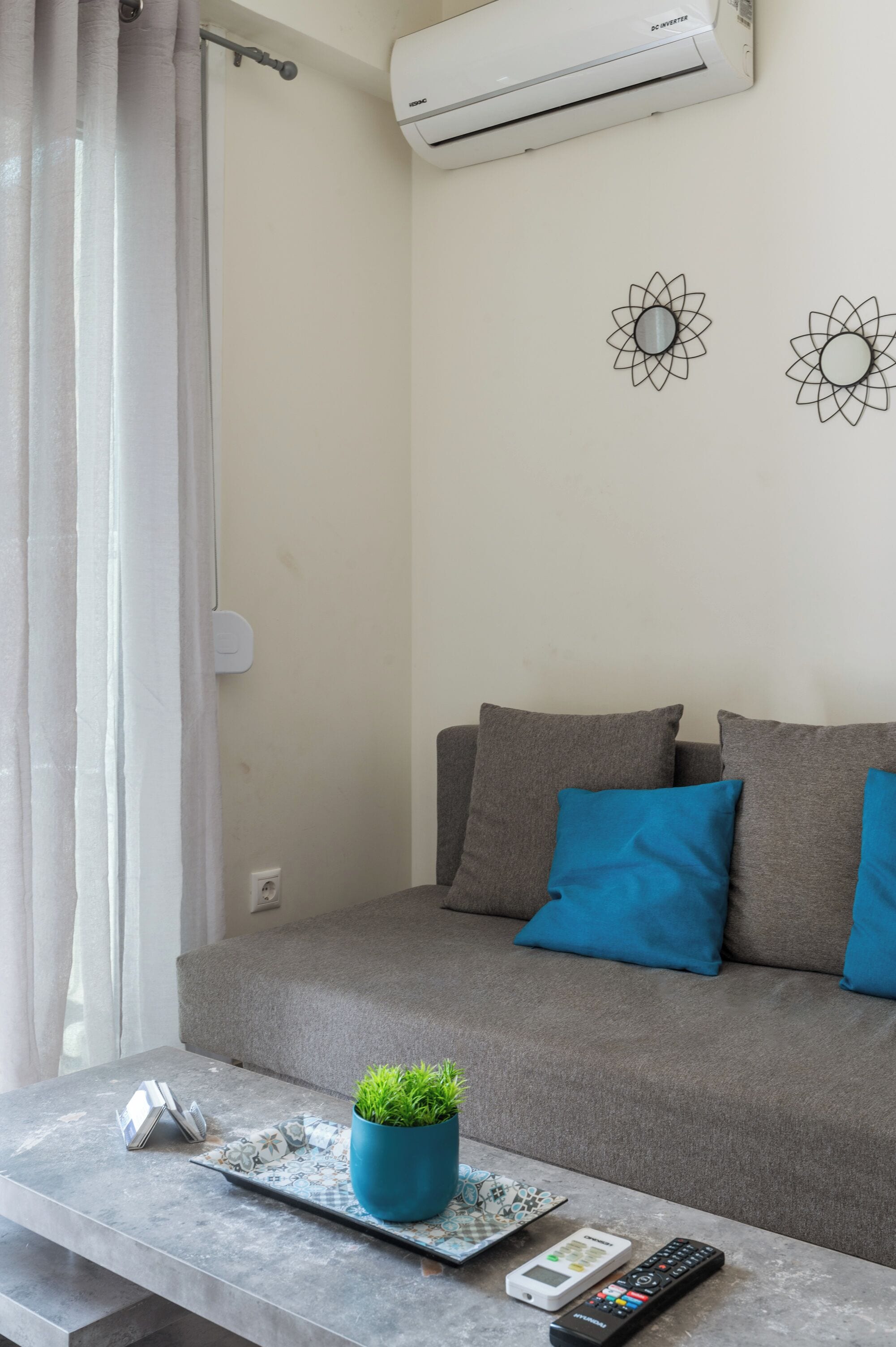 En stue med en grå sofa, blå puder, et lille bord med en plante, en fjernbetjening og dekorative billeder på væggen.