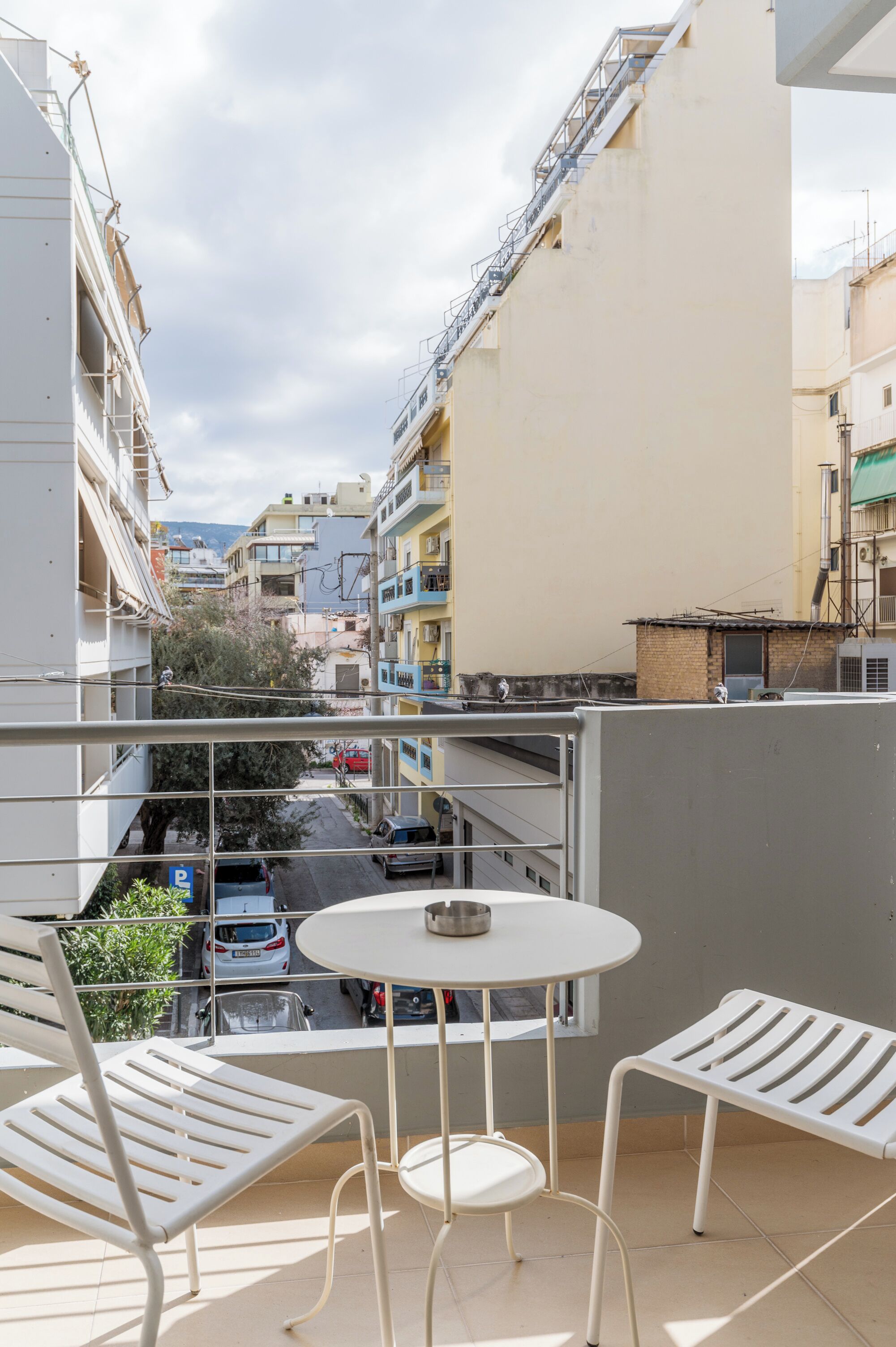 A balcony with a white table and chairs, overlooking a street with parked cars and buildings.