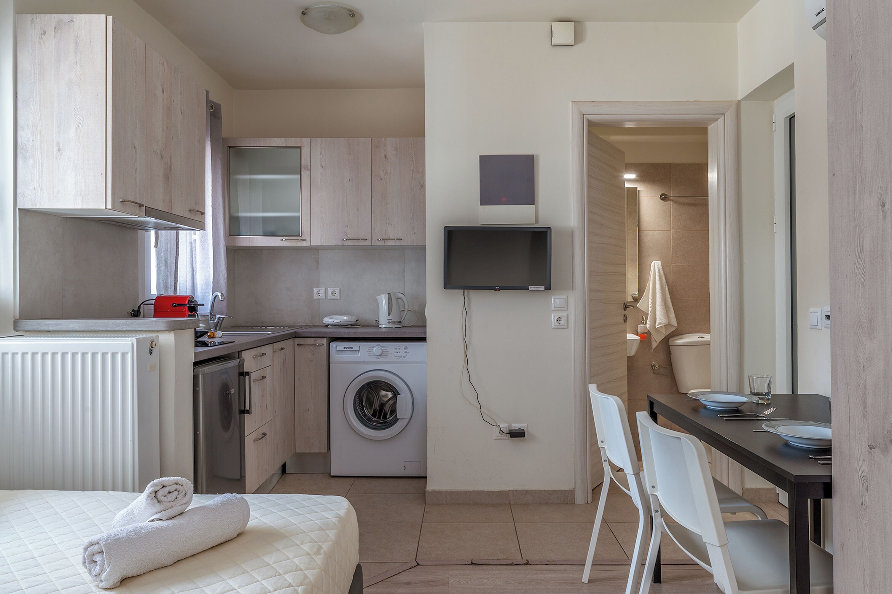 A compact kitchen with a washing machine, a dining area with chairs, and a bed with a rolled towel.