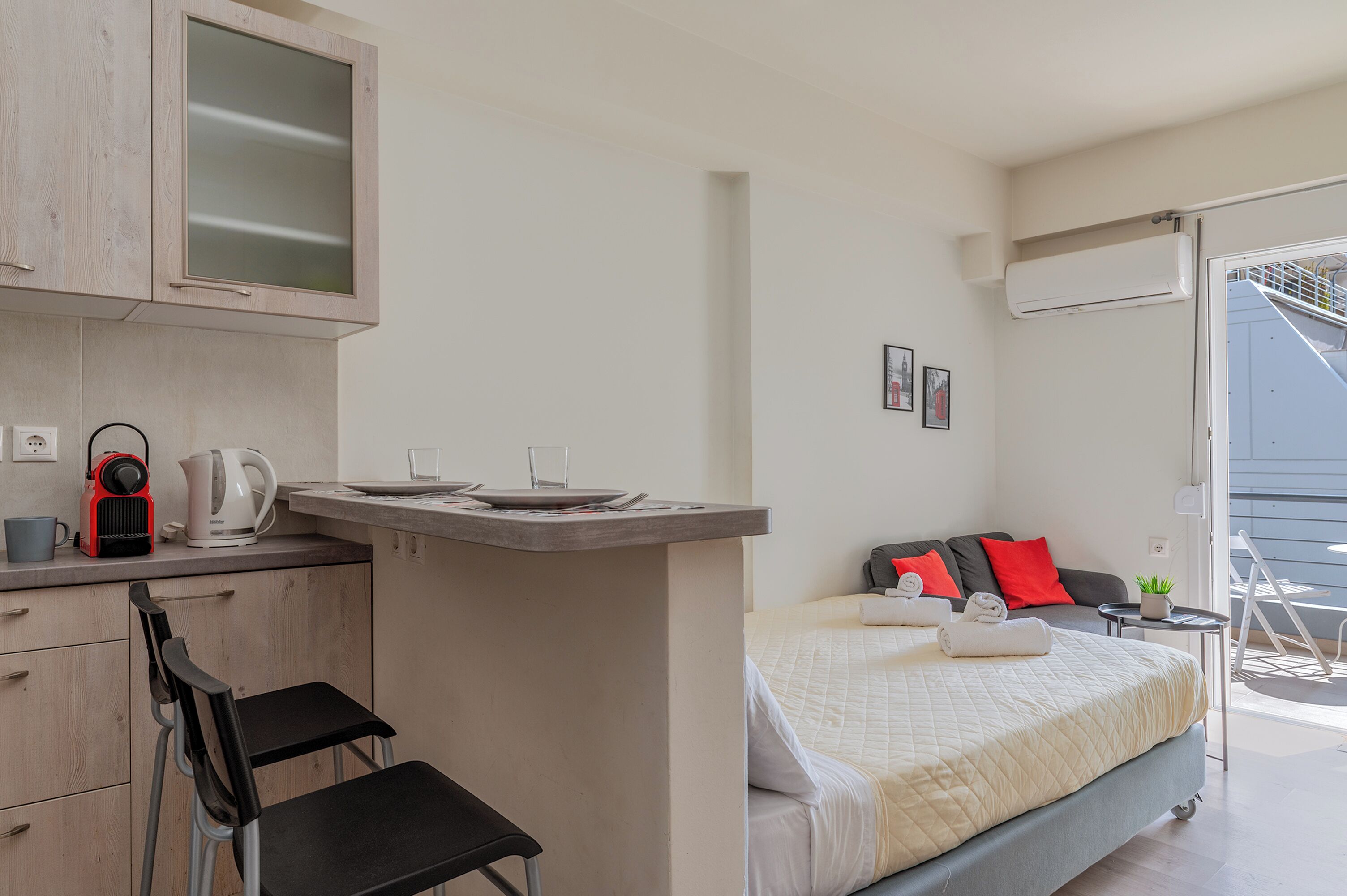 A compact hotel room with a kitchenette, a bed with red pillows, a small balcony, and a view of the outside.