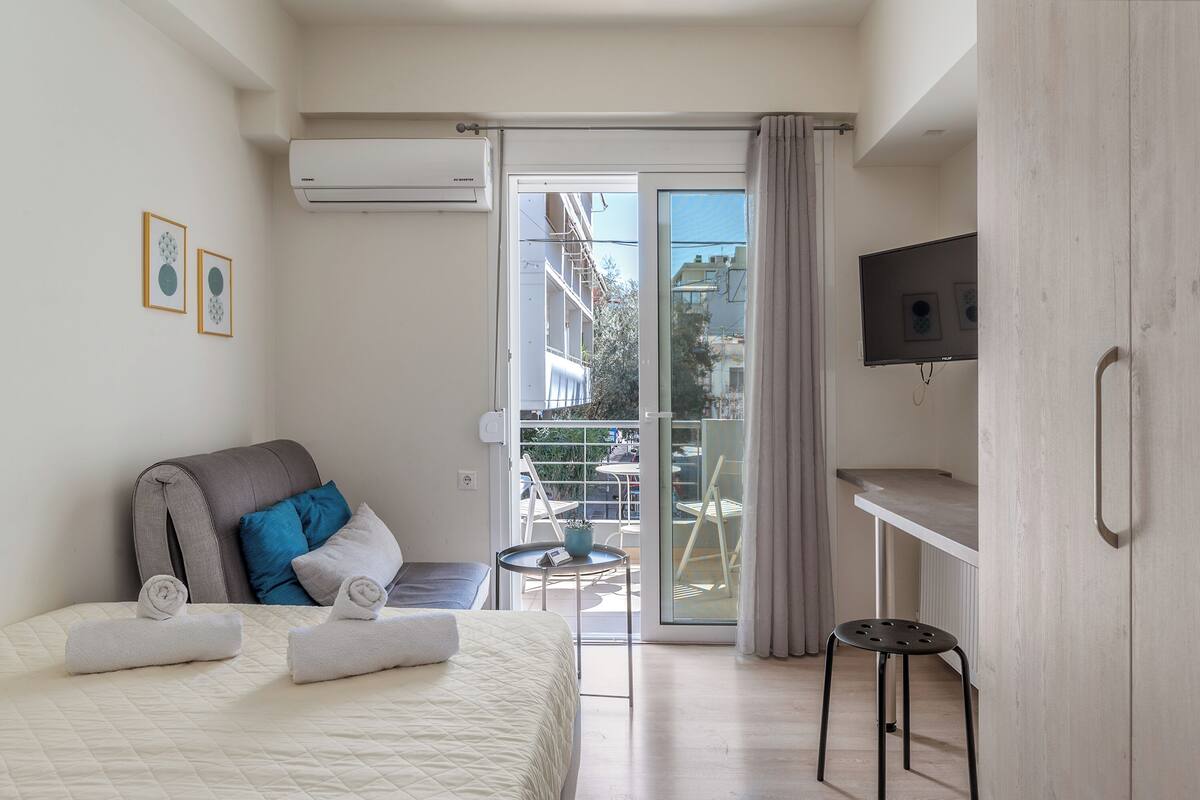 A modern hotel room with a bed, a small table, a chair, an air conditioning unit, and a balcony with a view of buildings.
