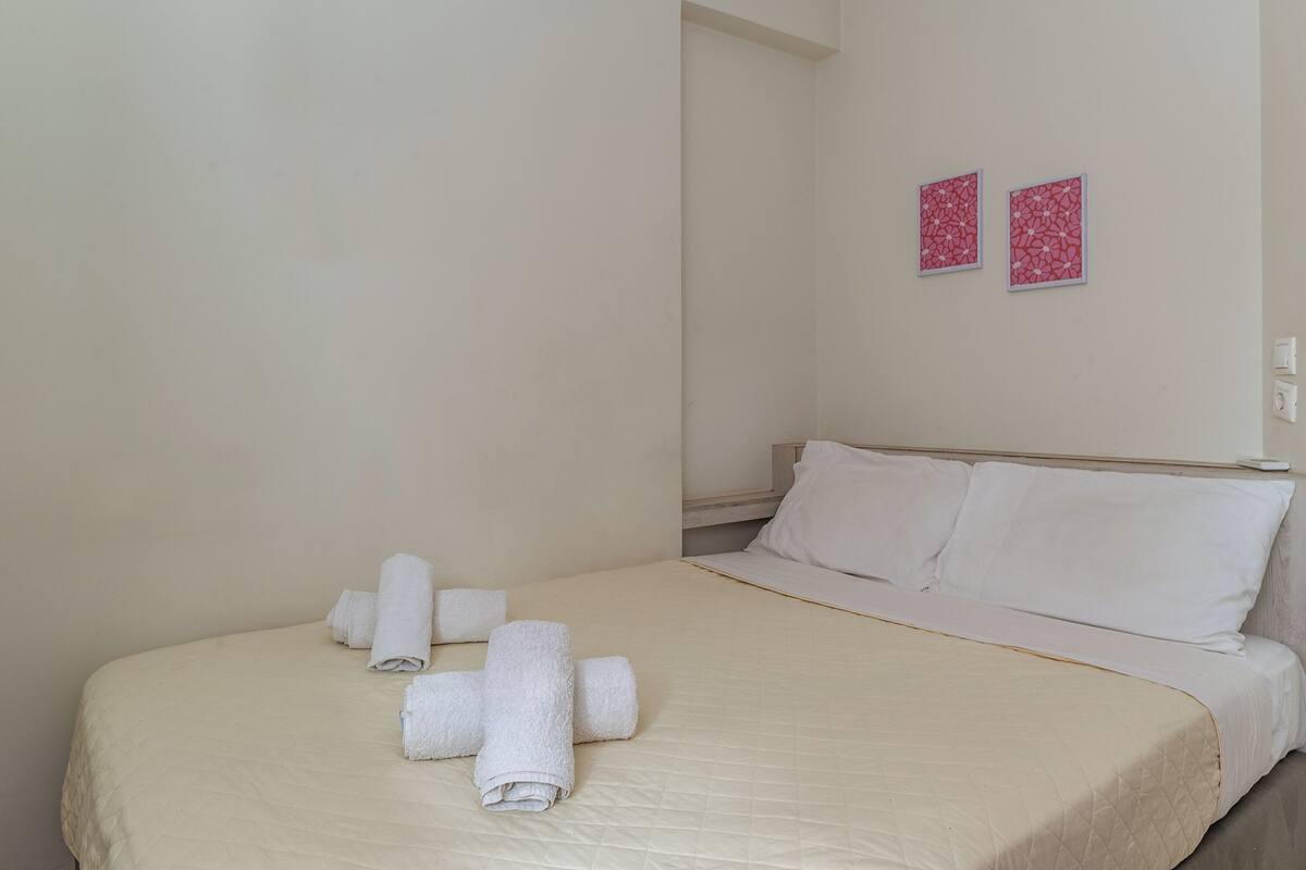 A neatly made bed with white linens and rolled towels, a plain wall with two framed pictures, and a light switch on the wall.