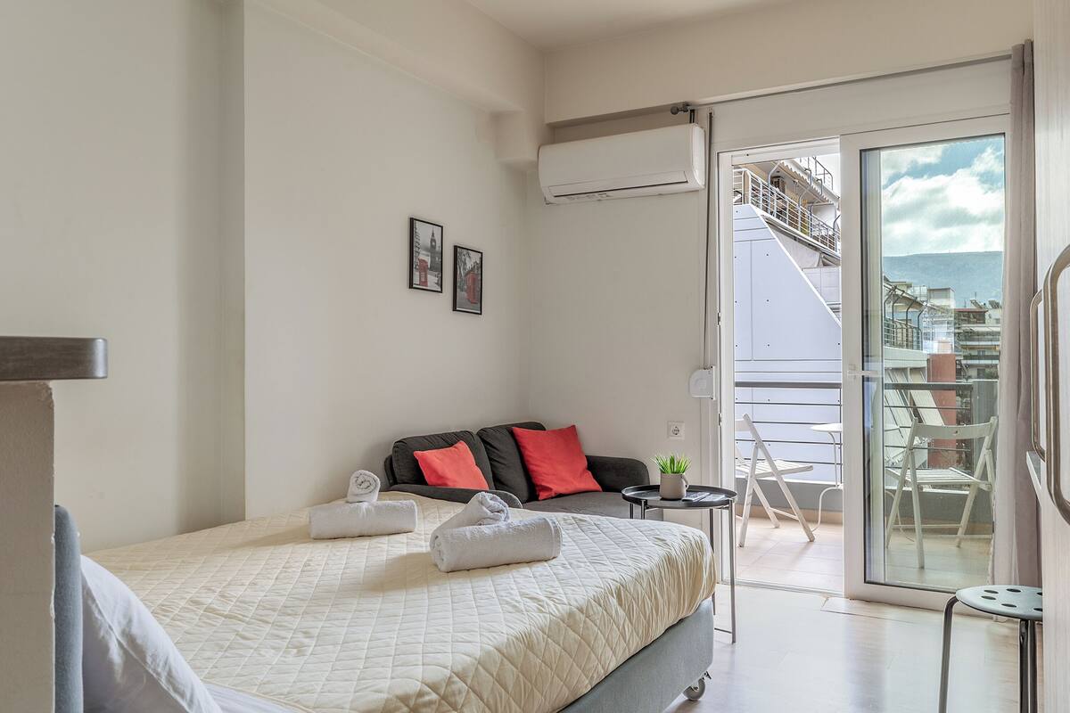 A hotel room with a bed, a sofa, a small table with a plant, and a balcony with chairs.