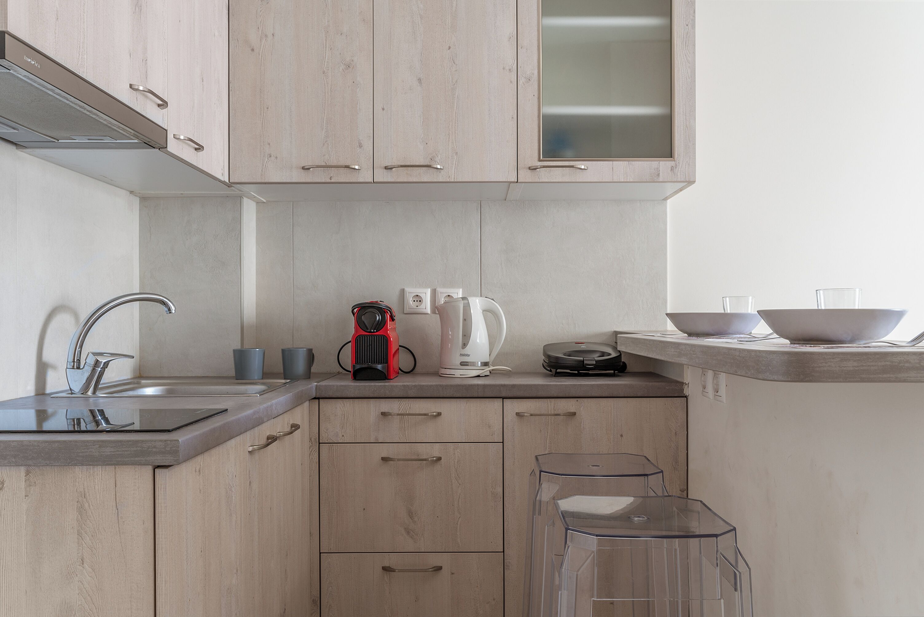 A modern kitchen with wooden cabinets, a stainless steel sink, a coffee maker, and a clear glass bar stool.