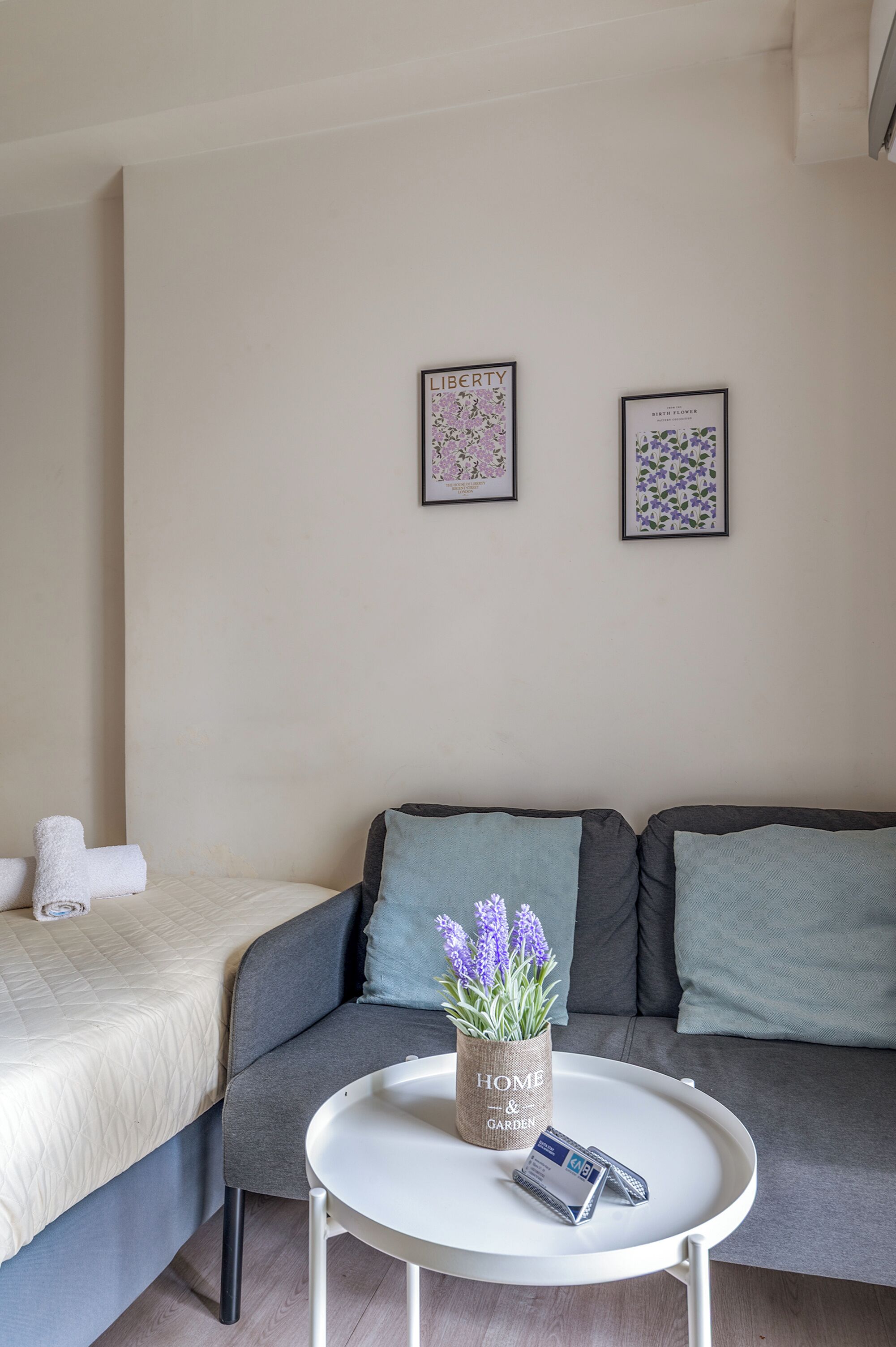 A modern bedroom with a bed, a grey sofa, a round white table, and two framed floral artworks on the wall.
