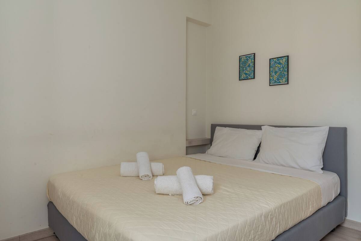 A neatly made bed with rolled white towels, a grey headboard, and two framed artworks on the wall.