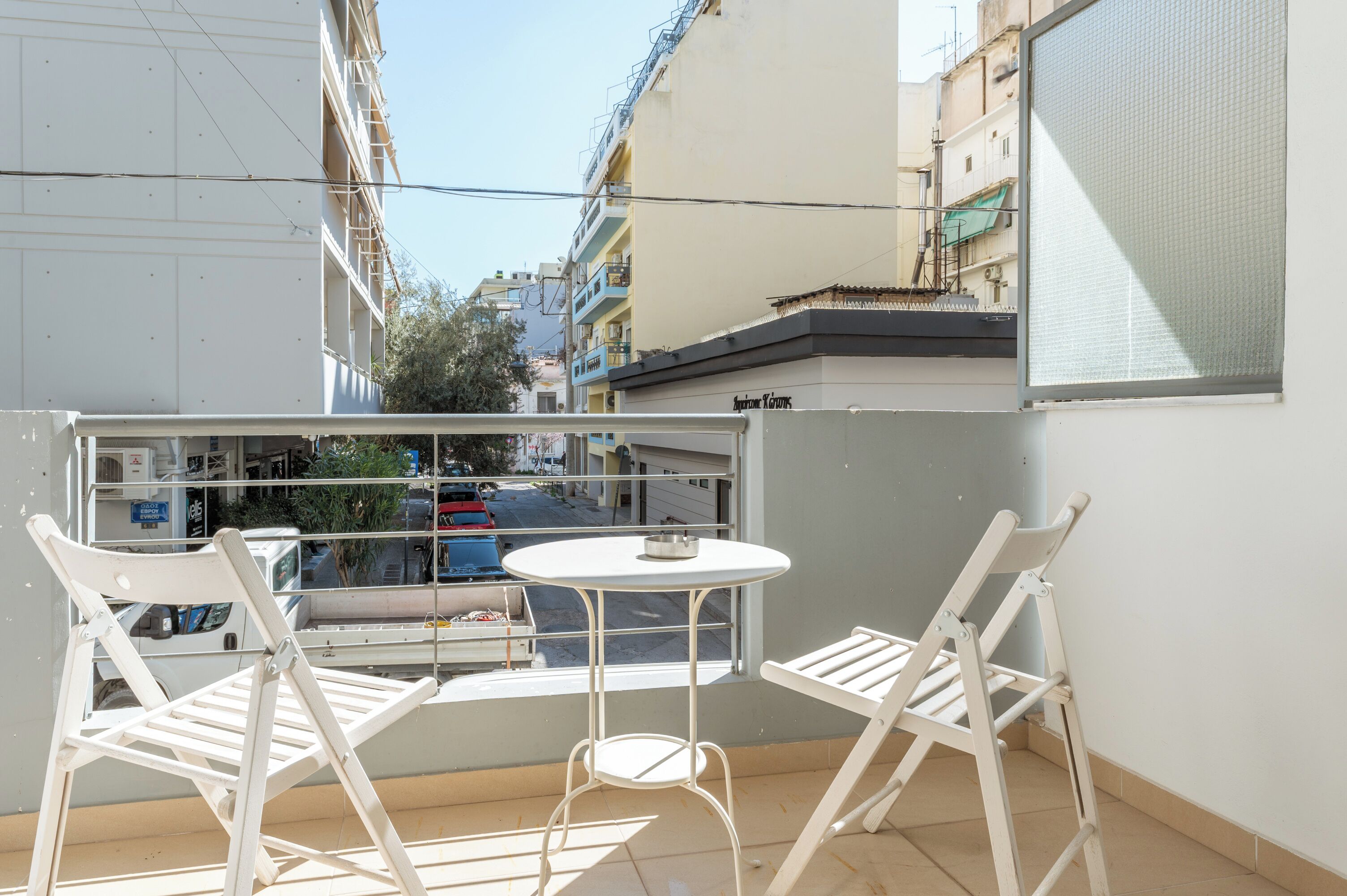 Un balcon avec une petite table et deux chaises blanches, donnant sur une rue bordée d’immeubles et de voitures garées.