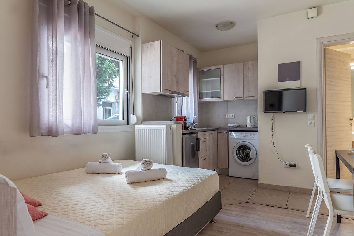 A modern hotel room with a bed, a small kitchenette, a washing machine, and a TV mounted on the wall.