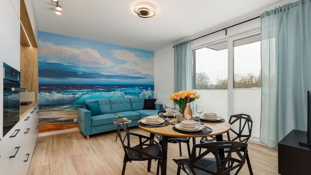 Ein modernes Wohnzimmer mit einem großen Meeresbild, einem blauen Sofa, einem Esstisch gedeckt mit Tellern und Gläsern sowie einem Fernseher auf einem Schrank.