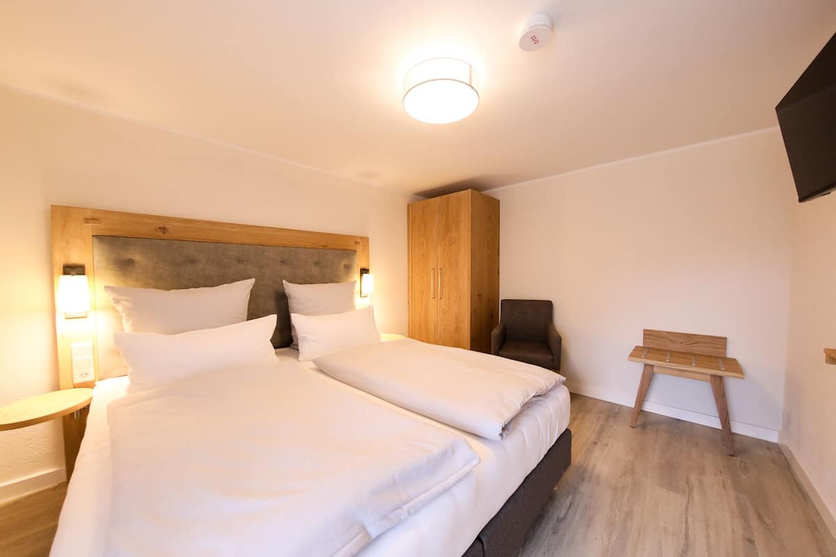 A modern hotel room with a large bed, a wooden headboard, a wardrobe, a small table, and a chair.