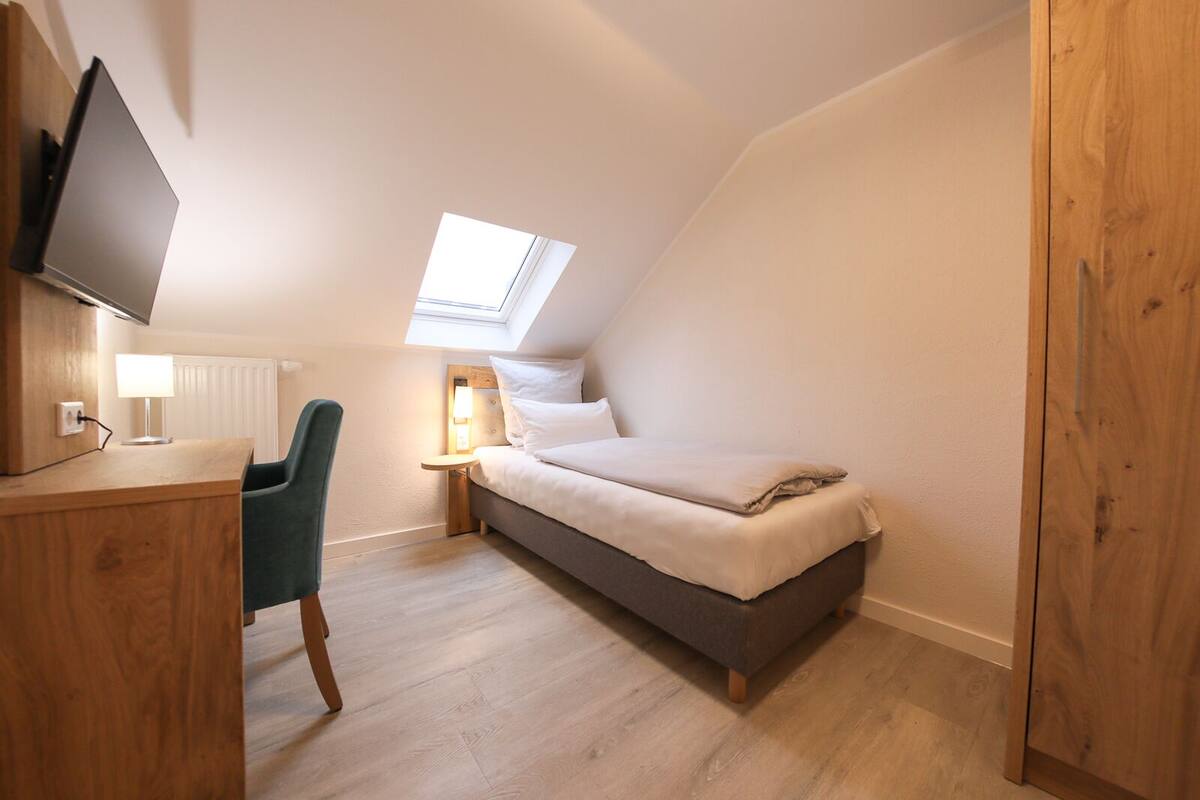 A compact bedroom with a bed, a desk, a chair, a bedside table with a lamp, and a skylight.