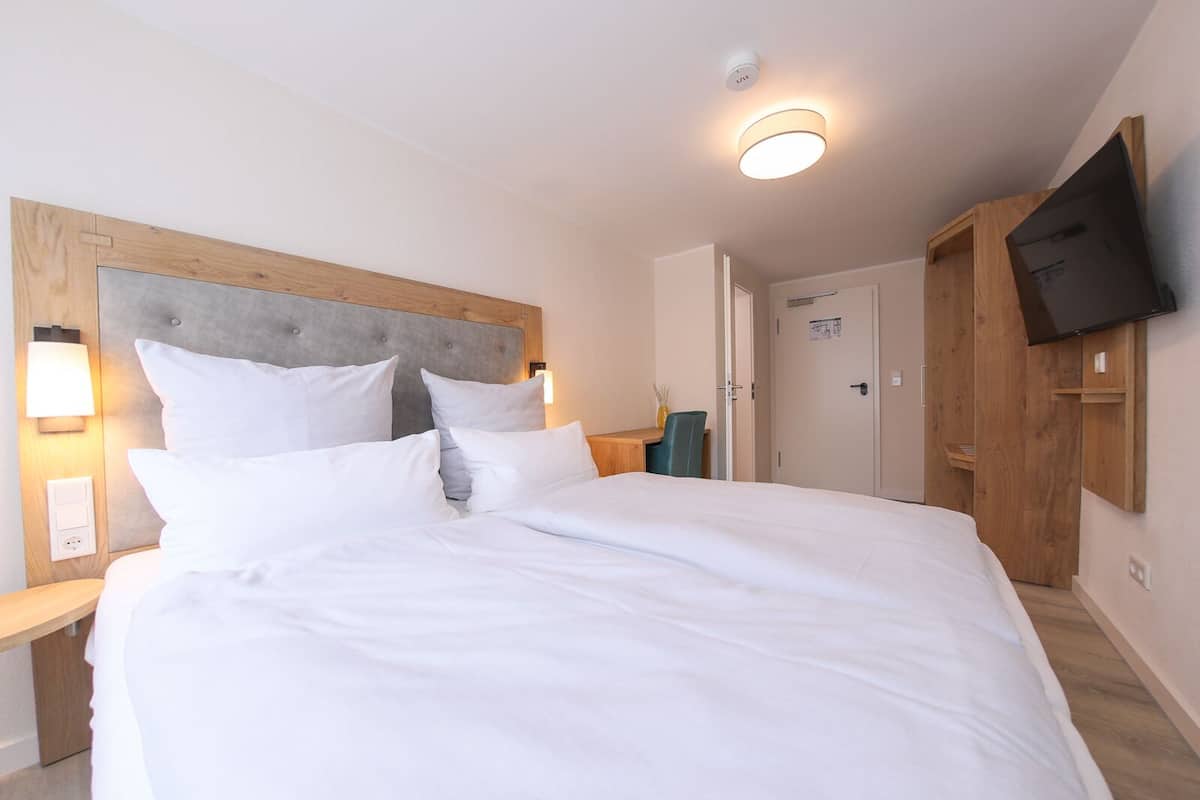 A hotel room with a large bed, wooden headboard, bedside lamps, a flat-screen TV mounted on the wall, and a door leading to another room.