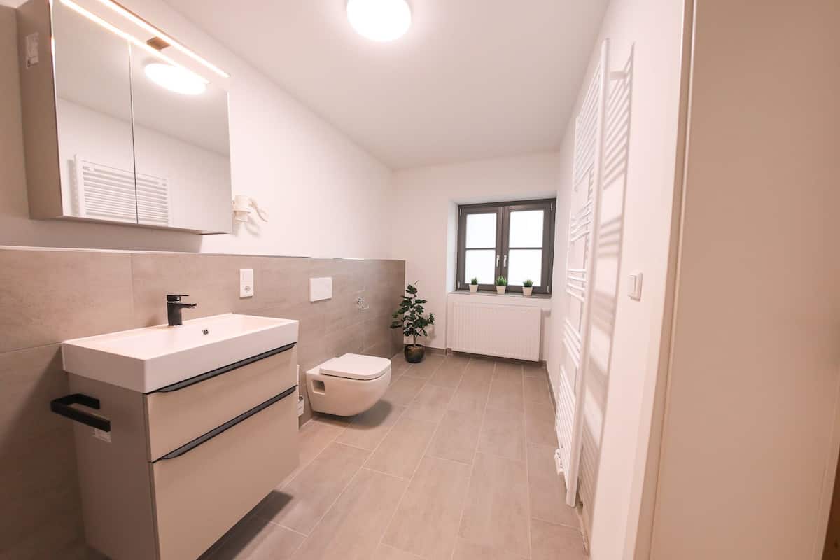 A modern bathroom with a sink, toilet, and a mirror. There is a window with plants on the windowsill and a radiator on the wall.