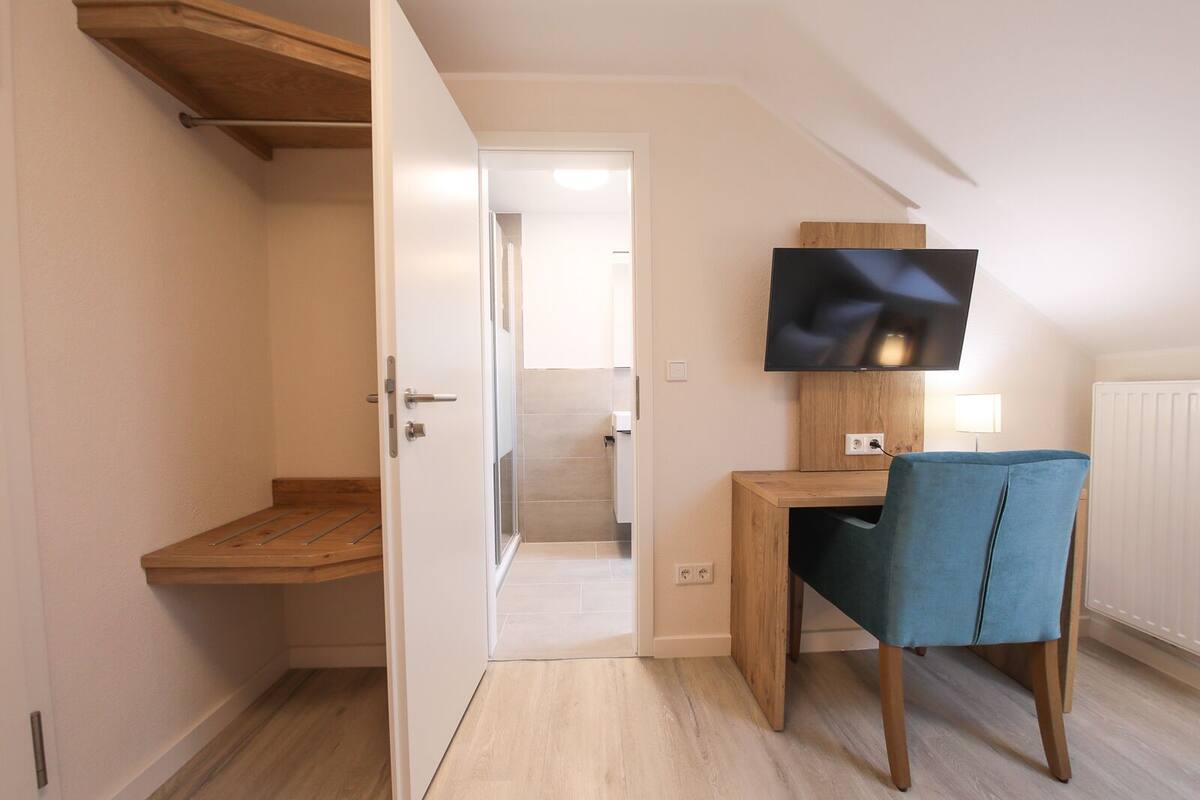 A modern room with a wooden desk, a blue chair, a flat-screen TV mounted on the wall, and a wooden shelf.