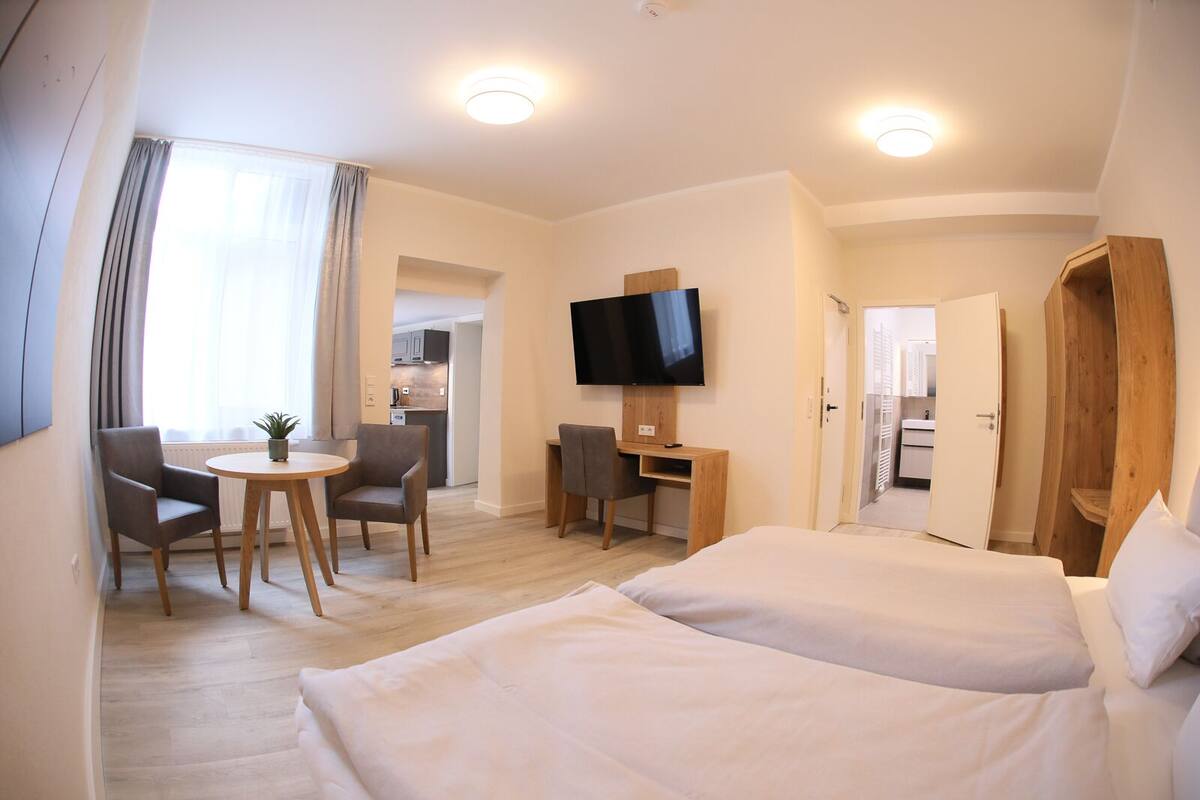 A hotel room with two beds, a desk, a TV, and a dining area with chairs and a table.