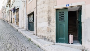 A narrow cobblestone street with buildings on either side. A green door with a number '4' sign is visible, and a suitcase is placed outside. - Eighteen 21 Houses - Casa de São Paulo (Lisboa)
