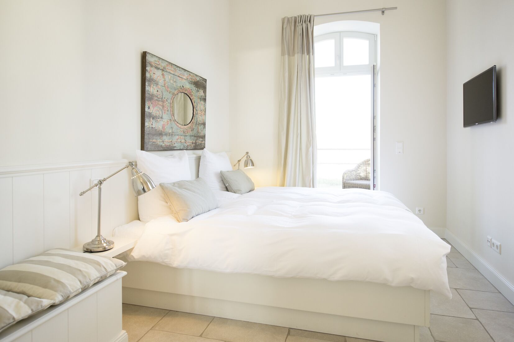 A modern bedroom with a large bed, a bedside table with a lamp, a window with curtains, and a wall-mounted TV.