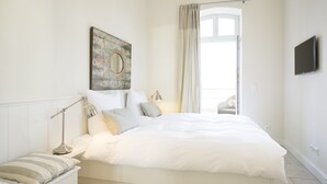 A modern bedroom with a large bed, a bedside table with a lamp, a window with curtains, and a wall-mounted TV. - Berliner Hof, Apartment 6 (55sqm), Westerland, Sylt. (Sylt)
