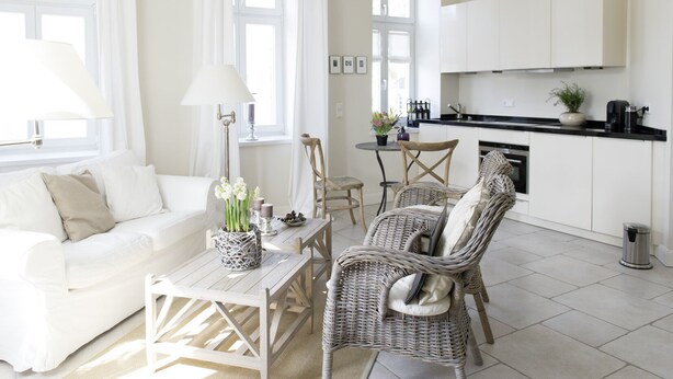 A modern, well-lit living room with a white sofa, wicker chairs, a coffee table with a vase of flowers, and a kitchen area with white cabinets and a black countertop. - Berliner Hof, Apartment 6 (55sqm), Westerland, Sylt. (Sylt)