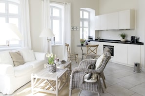 A modern, well-lit living room with a white sofa, wicker chairs, a coffee table with a vase of flowers, and a kitchen area with white cabinets and a black countertop. - Berliner Hof, Apartment 6 (55sqm), Westerland, Sylt. (Sylt)