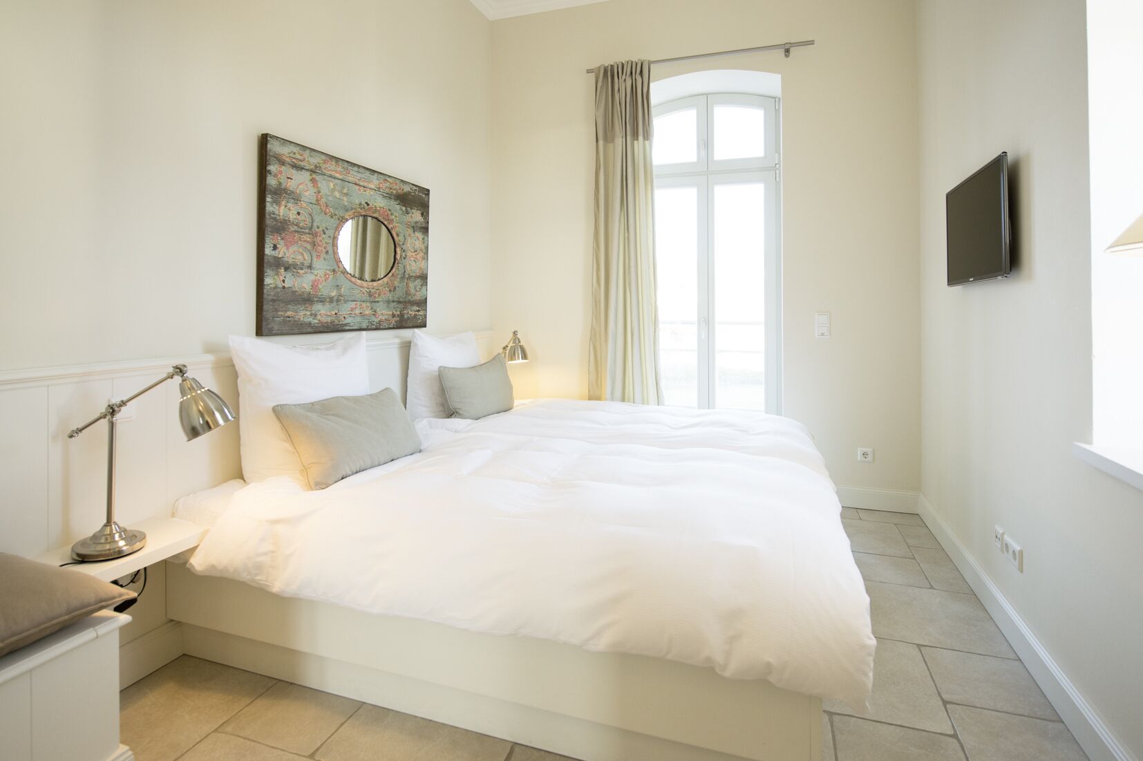Une chambre à coucher avec un grand lit, une table de chevet avec une lampe, un téléviseur fixé au mur et une fenêtre avec des rideaux.