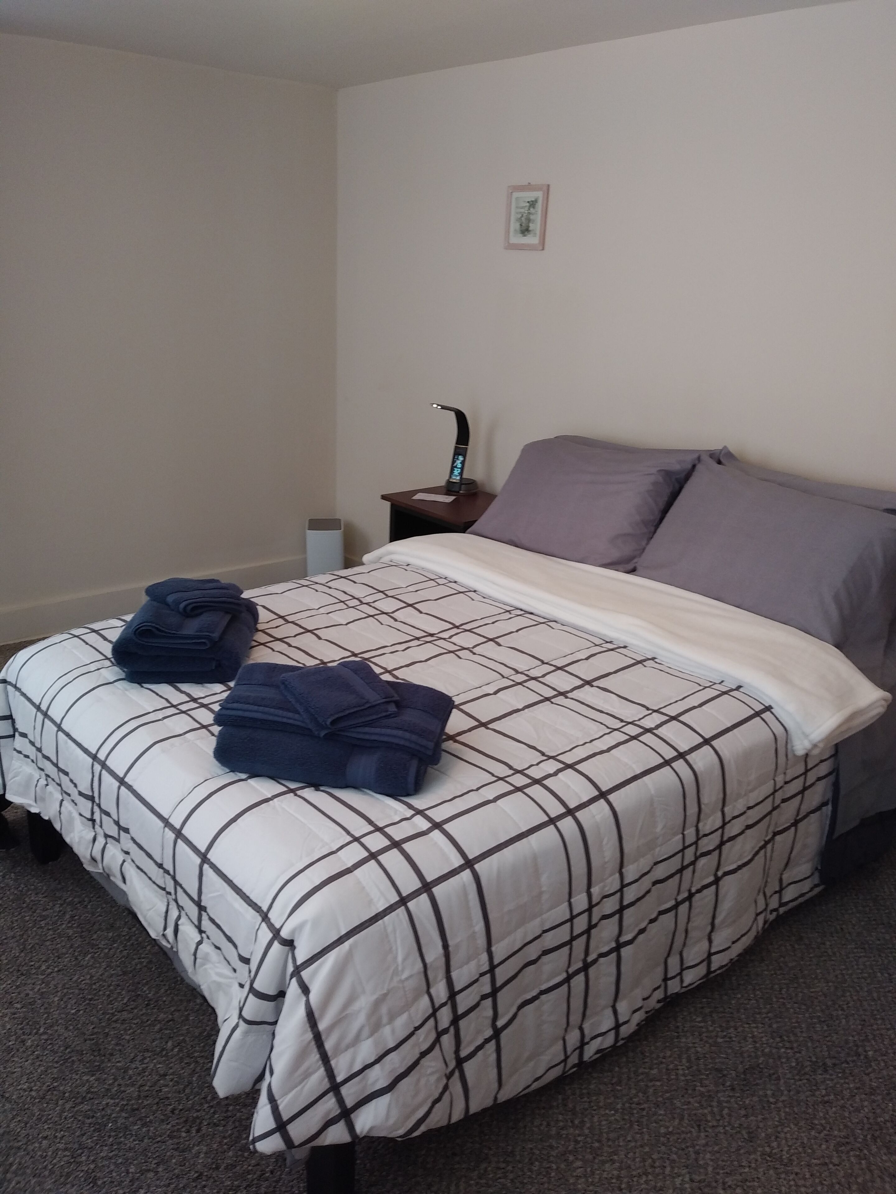A neatly made bed with a checkered bedspread, two folded towels, and a nightstand with a lamp.