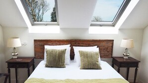 Une chambre à coucher avec une tête de lit en bois, deux fenêtres de toit, un lit avec du linge de lit blanc, deux tables de chevet avec des lampes et une vue sur les arbres à travers les fenêtres de toit.