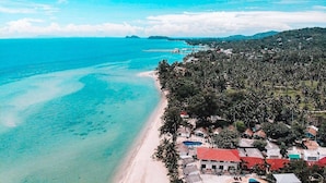 Vista aérea de uma área costeira com praia, piscina e um conjunto de edifícios com telhados vermelhos.