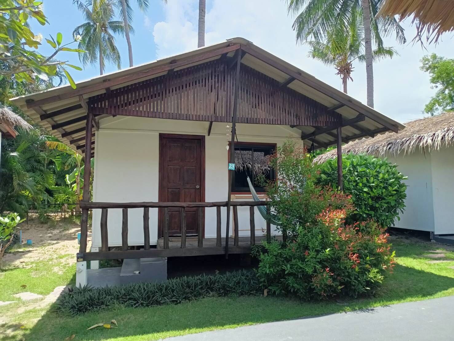 A small, single-story house with a thatched roof, surrounded by lush greenery and palm trees.
