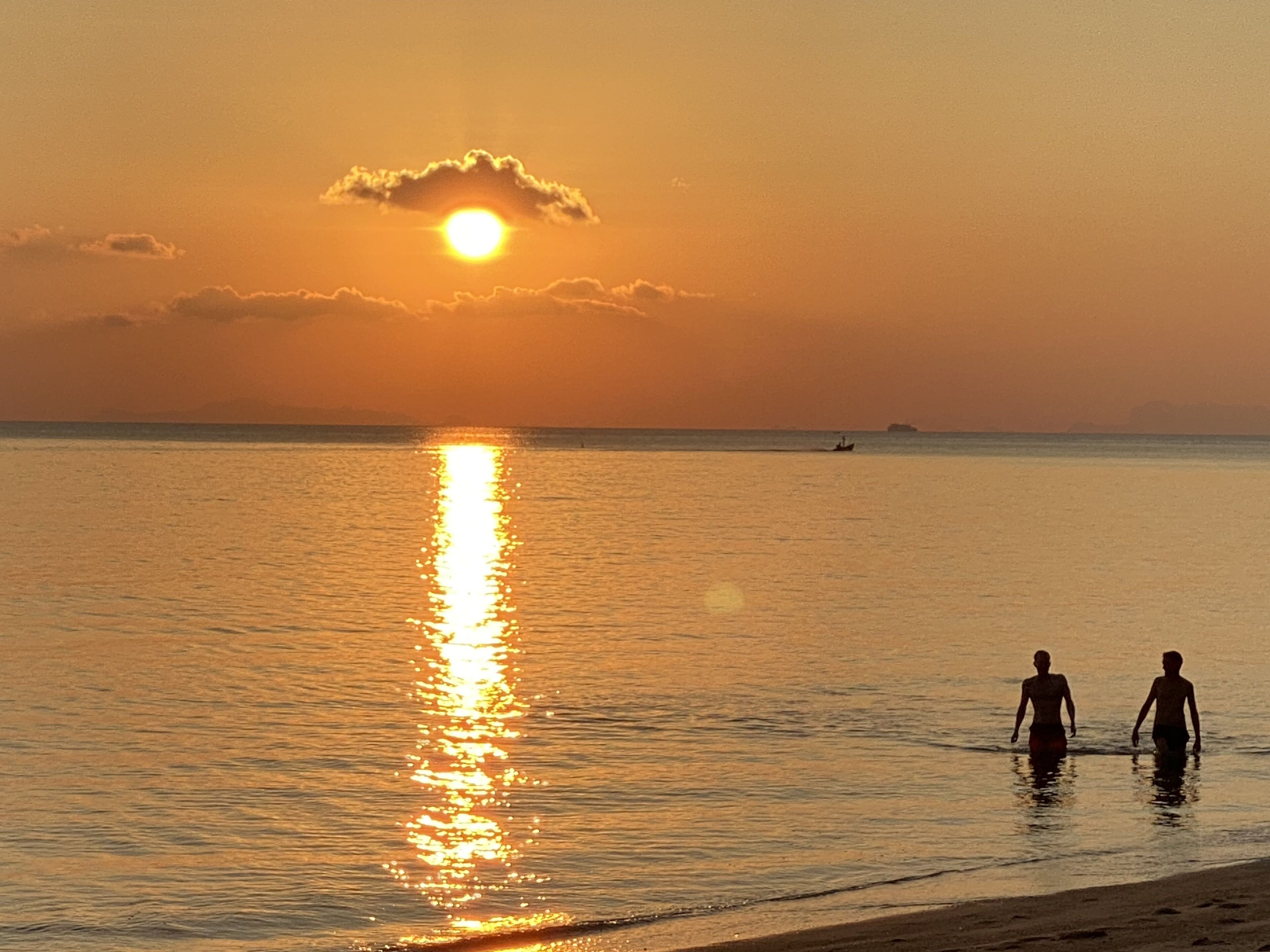A beach scene with two people walking in shallow water, a boat in the distance, and a sunset over the ocean.