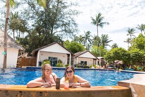 A swimming pool area with two people relaxing by the poolside, surrounded by tropical vegetation and thatched-roof structures.