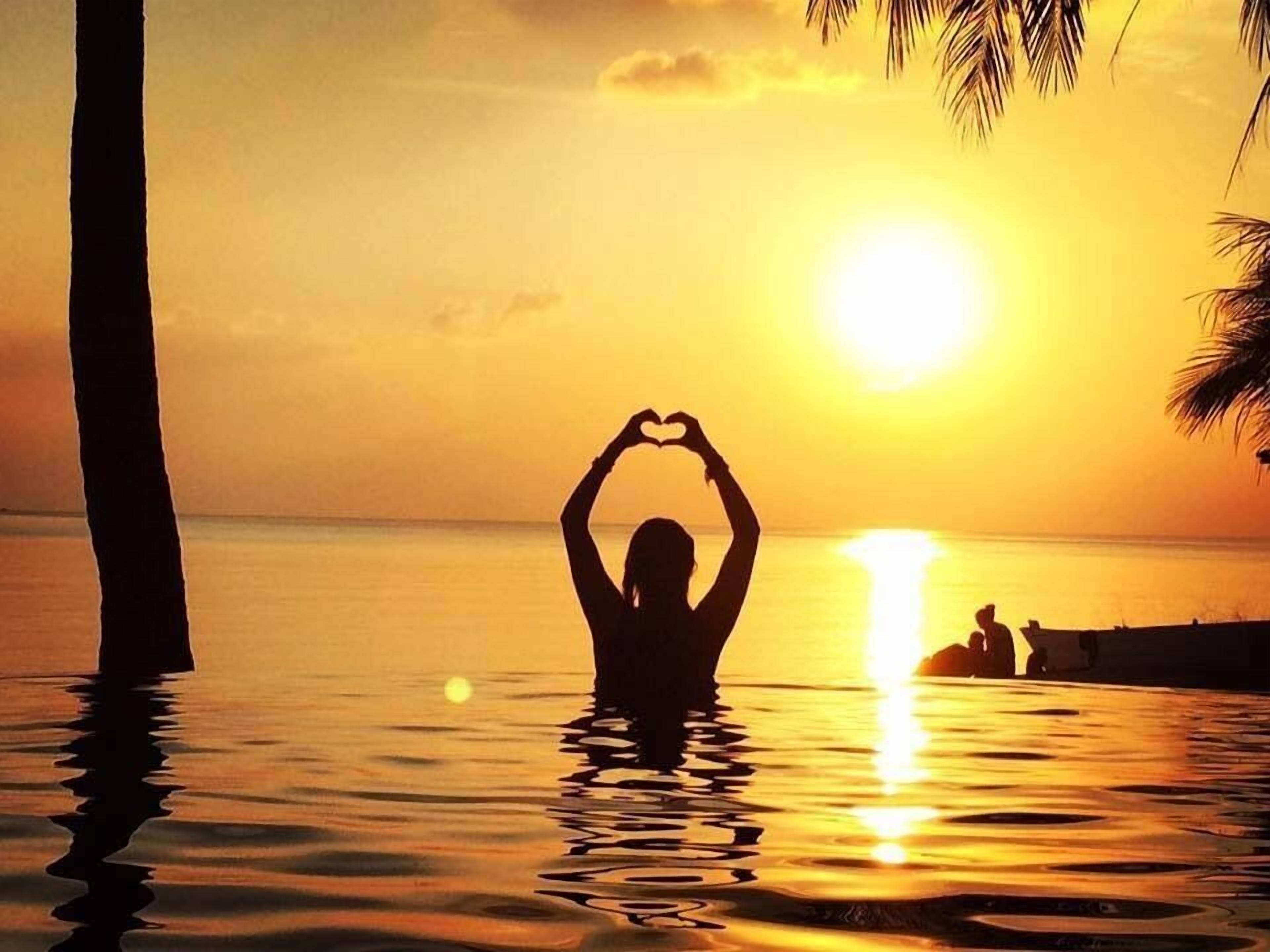 A person making a heart shape with their hands in the water at sunset.