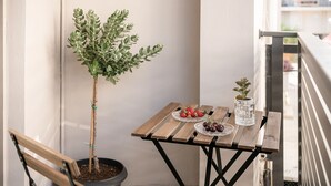 Un banco de madera, una mesa pequeña con una planta en maceta, un vaso de agua y un plato de fresas y uvas en un balcón.