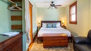 A bedroom with a bed, a wooden dresser, a chest of drawers, a ceiling fan, a nightstand, a lamp, and a window with blinds.