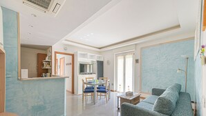 A modern living room with a grey sofa, a dining area with blue chairs, a kitchen with a fridge and microwave, and a large window with curtains. - Norb House Lerici ( CIN IT011016C2Y3WHZASD) (Lerici)
