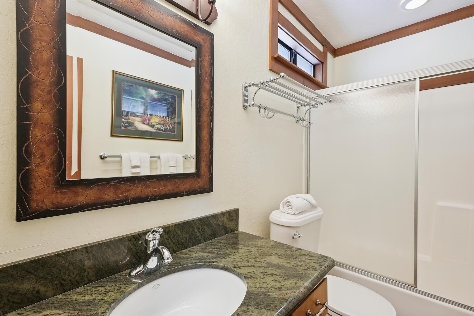 A bathroom with a granite countertop, a sink, a mirror, a framed picture, a towel rack, a toilet, and a shower.