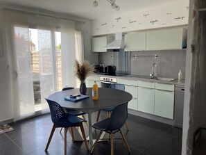 A modern kitchen with a dining area, featuring a round table, two chairs, a vase with dried plants, and a bottle of orange juice.