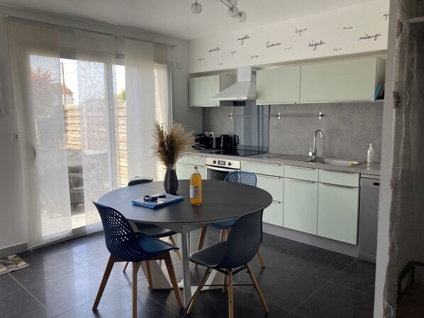 A modern kitchen with a dining area, featuring a round table, two chairs, a vase with dried plants, and a bottle of orange juice.
