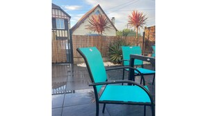Een terras buiten met twee turquoise stoelen en een tafel, omringd door een bamboomborduurwerk en een huis op de achtergrond.