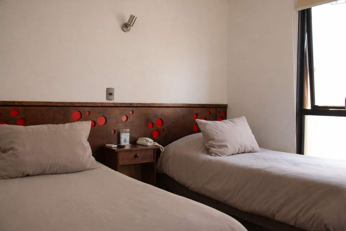 Camera d'albergo con due letti, una testiera in legno con motivi circolari rossi, un comodino con un telefono e una lampada, e una finestra che lascia entrare luce naturale.