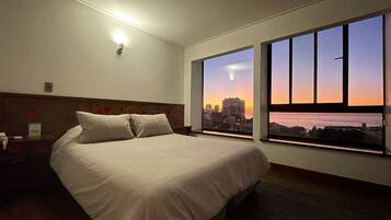 A hotel room with a large bed, a nightstand, a wall-mounted light, and a window with a cityscape view.