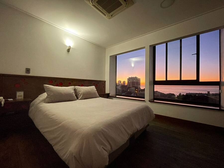 A hotel room with a large bed, a nightstand, a wall-mounted light, and a window with a cityscape view.