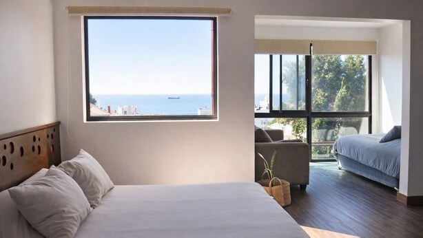 Un dormitorio con un ventanal que ofrece vistas al mar, una cama con almohadas, un sofá y una planta pequeña.