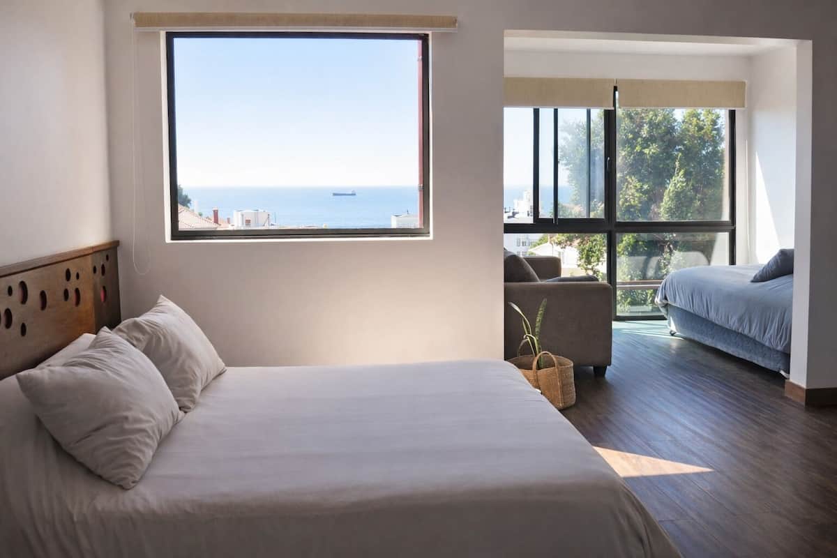 Camera da letto con un'ampia finestra vista mare, un letto con cuscini, un divano e una piccola pianta.