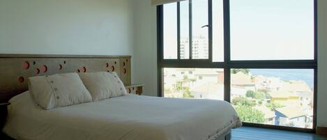 A bedroom with a large bed, a nightstand, and a window with a view of buildings and a body of water.